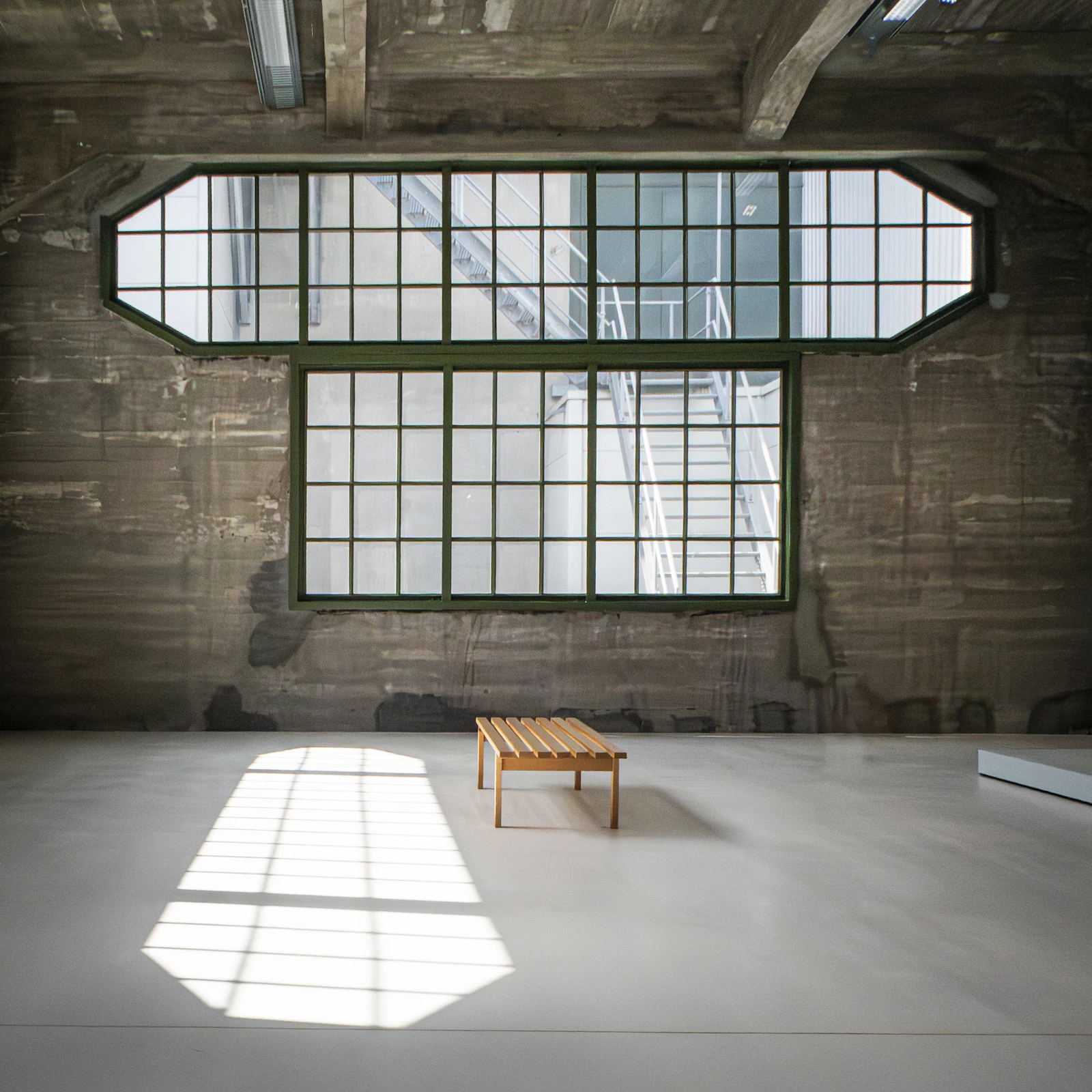 Sunlight through large industrial windows falls across a concrete floor and a single wooden bench
